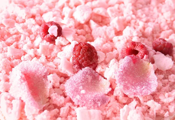 A closeup top view of a pink cake covered with meringue crumbles, raspberries, and sugary rose petals.