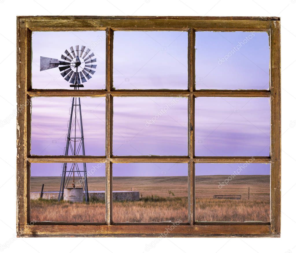 windmill with a pump and cattle water tank in shortgrass prairie at dusk as seen from a vintage cabin window