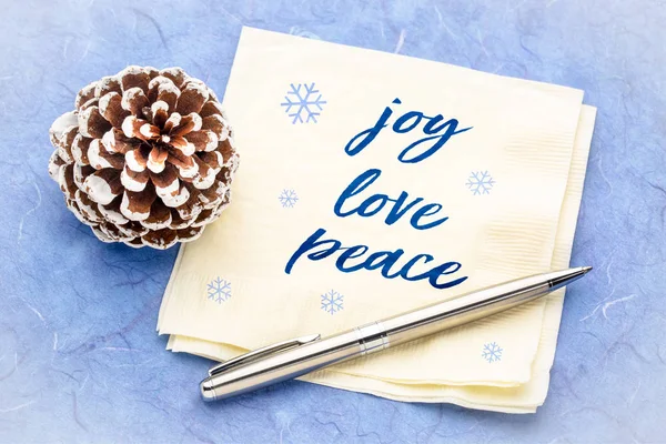 joy, love, peace concept - handwriting on a napkin with pine cone decoration against blue mulberry paper