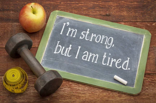 I am strong, but I am tired - white chalk text on a slate blackboard against weathered red painted barn wood with a dumbbell, apple and tape measure