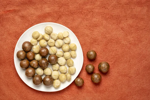 Tuercas Macadamia Plato Blanco Contra Fondo Papel Texturizado Rojo Con — Foto de Stock