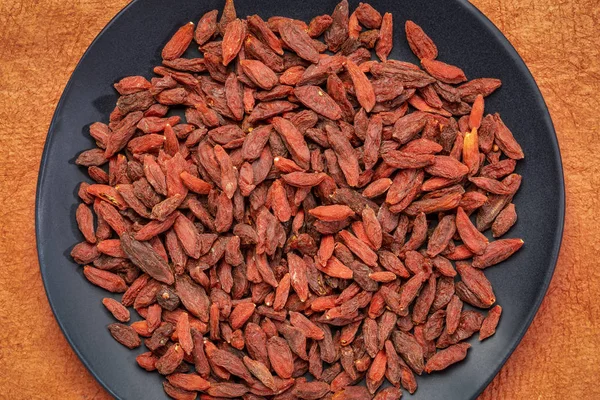 Dried Goji Berries Black Ceramic Plate Orange Textured Paper Background — Stock Photo, Image