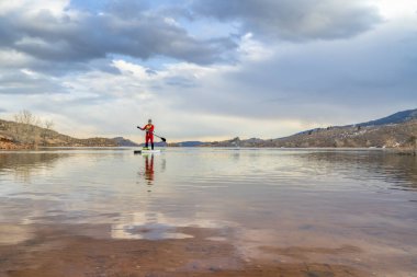 senior male paddler in a dry suit and life jacket is paddling a stand up paddleboard on a mountain lake in winter conditions - Horsetooth Reservoir in northern Colorado clipart