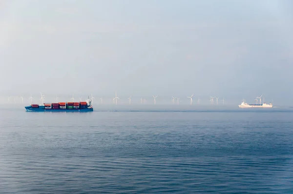 Konteynır gemileri sabah sisinde kıyıdan uzak bir rüzgar parkının önünden geçiyor. — Stok fotoğraf