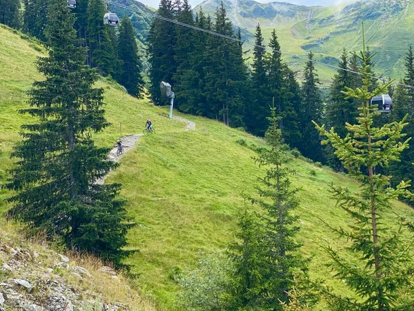 Motociclistas irreconocibles en un sendero de bicicleta de montaña en Saalbach en los Alpes austríacos en verano con cielo dramático y teleférico en el fondo — Foto de Stock