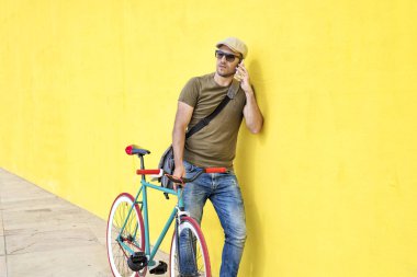 Vintage bisiklet ve rahat kıyafetler ve güneşli bir günde bir cep telefonu kullanırken sarı bir duvara karşı duran güneş gözlüğü takan genç bir adamla yan görünüm