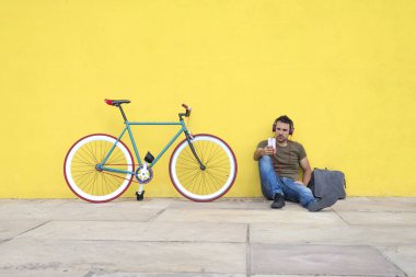 Sarı bir duvara ekranında bakarken kulaklıklar sahip bir cep telefonu kullanarak trendy bakışla katta oturan çekici bir erkek önden görünümü