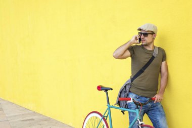 Vintage bisiklet ve rahat kıyafetler ve güneşli bir günde bir cep telefonu kullanırken sarı bir duvara karşı duran güneş gözlüğü takan genç bir adamla yan görünüm