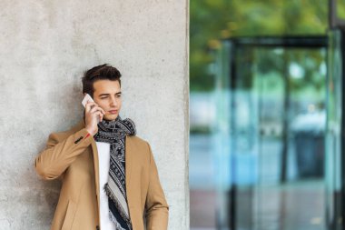 Cep telefonu açık havada kullanırken bir duvara yaslanmış kot kıyafetleri şık genç adam önden görünümü