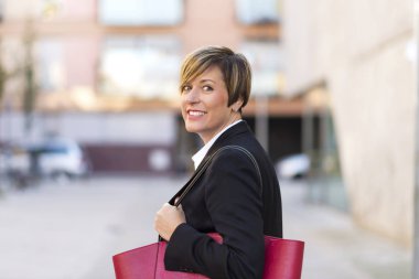 Güneşli bir günde kamera açık havada bakarken Caddesi üzerinde duran bir güven, zarif ve gülümseyen iş kadını arkadan görünüş