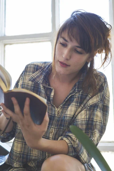 Low angle view of a beautiful sensual woman wearing shirt with ponytail sitting on staircase at home while holding a book next to a window in a sunny day