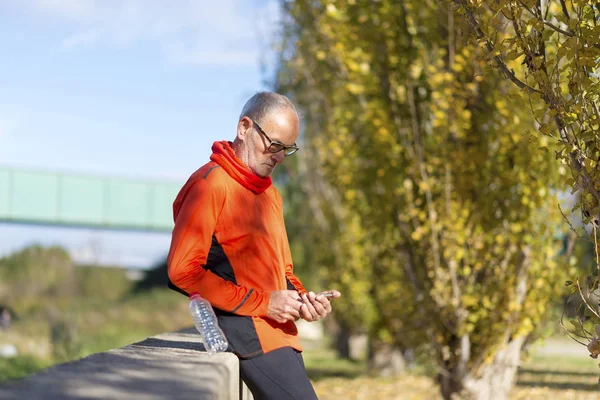 Side view of a senior runner man leaning on fence while testing exercise in a mobile phone outdoors in a sunny day — Stock Photo, Image