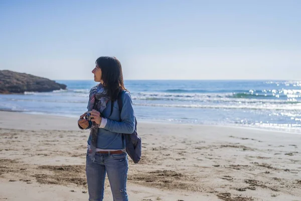 Вид спереди на молодую женщину в повседневной одежде, стоящую на пляже, держа камеру и глядя в яркий день — стоковое фото