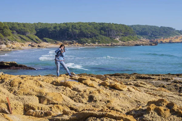 Front view of young woman wearing casual clothes standing on seashore while taking a photograph in a bright day — Stock Photo, Image