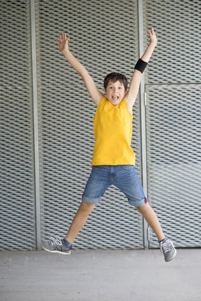 Front view of a smiling cheerful boy jumping while arms raised outdoors in a bright day — Stock Photo, Image