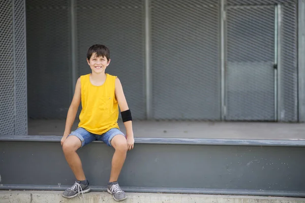 明るい日にカメラを見ながら金属製の柵の上に座っている笑顔の少年の正面図 — ストック写真