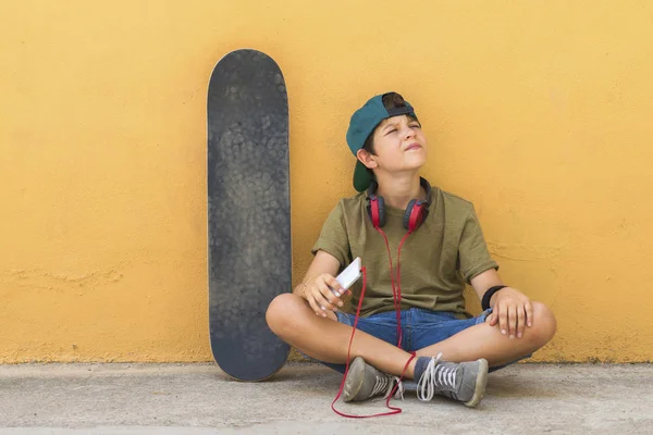 Front bild av ung pojke som sitter på marken lutar på en gul vägg medan du använder en mobiltelefon för att lyssna på musik — Stockfoto