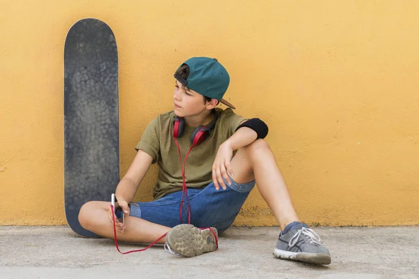 Front view of young boy sitting on ground leaning on a yellow wall while using a mobile phone to listening music — Stock Photo, Image