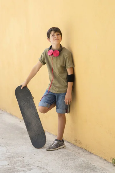 Young boy leaning on a yellow wall with headphones on neck, holding a skateboard while looking away in a bright day — Stock Photo, Image