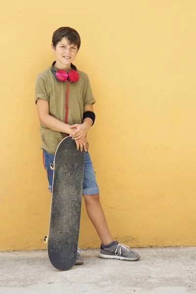 Young boy leaning on a yellow wall with headphones on neck, holding a skateboard while looking away in a bright day — 스톡 사진