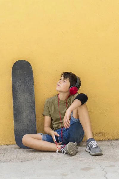 Vista frontal de um jovem patinador sentado contra uma parede amarela enquanto ouve música por fones de ouvido — Fotografia de Stock