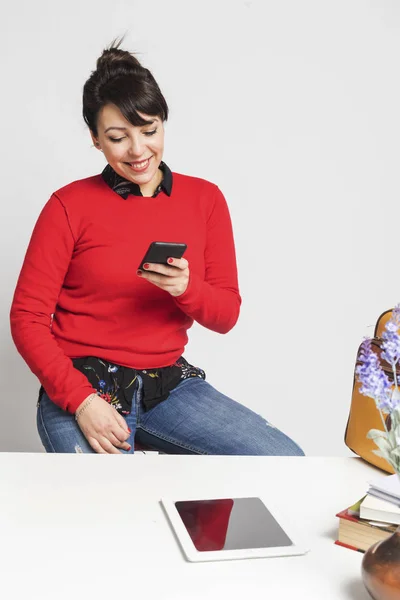 Вид спереди красивой латиноамериканской женщины, сидящей на табуретке во время использования мобильного телефона в офисе на белом фоне — стоковое фото