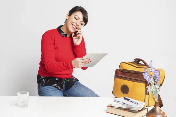 Вид спереди красивой женщины с пончиком, сидящей на табуретке, пользуясь телефоном и держа планшетный компьютер на белом фоне — стоковое фото