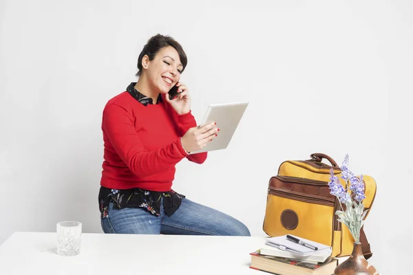 Vista frontal de uma mulher bonita com rabo de cavalo sentado em um banquinho enquanto usa o telefone e segurando um tablet pc contra fundo branco — Fotografia de Stock