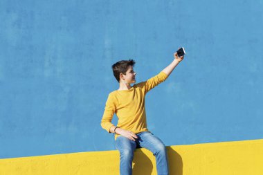 Akıllı telefonla selfie çekerken mavi duvara karşı sarı bir çitin üzerinde oturan rahat kıyafetler giyen genç bir çocuğun ön görünümü
