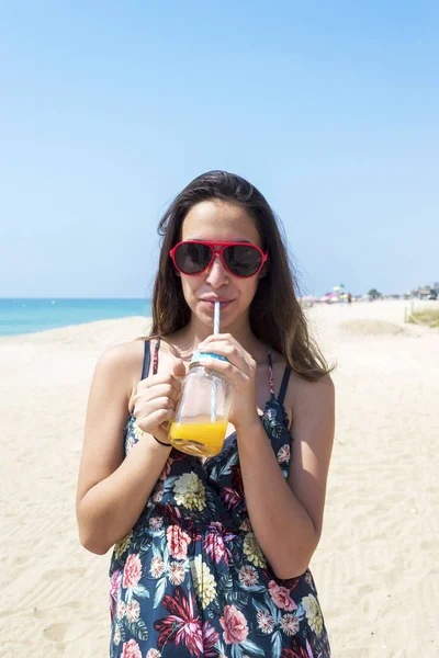 Ein junges braungebranntes Mädchen kam in das Resort, trägt ein modisches Kleid und eine Sonnenbrille, hält ein leckeres Saftgetränk in der Hand. — Stockfoto