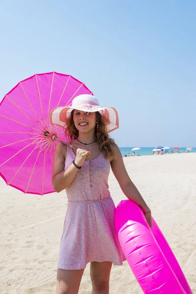 Mulher bonita em vestido rosa andando na praia de areia usando um chapéu e segurando um guarda-chuva e um flutuador — Fotografia de Stock