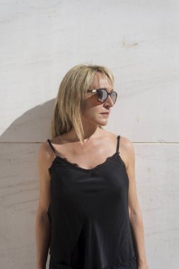 Sarışın kadın, beyaz bir duvara yaslanmış, başka tarafa bakıyor.