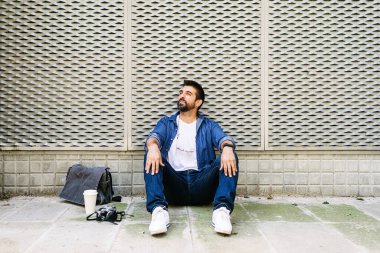 Keyifli sakallı adam, günlük giysiler içinde sokak zemininde oturmuş kahve molası verirken dinleniyor.