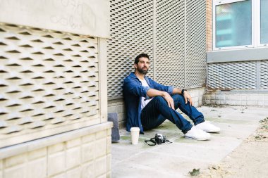 Keyifli sakallı adam, günlük giysiler içinde sokak zemininde oturmuş kahve molası verirken dinleniyor.