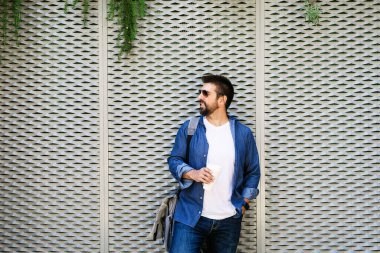 Mavi tişörtlü yakışıklı bir adamın portresi. Elinde kahveyle metalik bir arka planda duruyor.