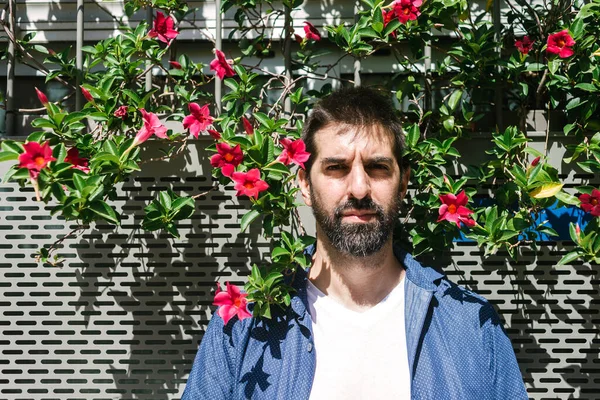 Bearded male in casual attire leaning on a flowered wall while looking camera