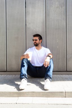 Güneş gözlüklü sakallı adam elinde kahve tutarken yerde oturuyor.