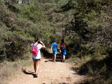 Sırt çantalı aile yolda yürüyor, anne çocuklara fotoğraf çekiyor.