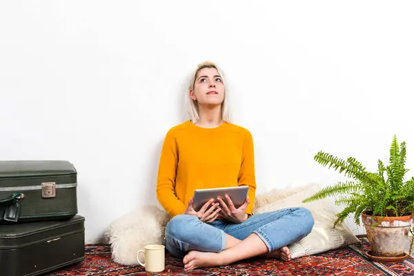 A pensive young woman with a tablet gazes upward, reflecting inspiration or curiosity, in a cozy home environment enhanced by plants and vintage elements.