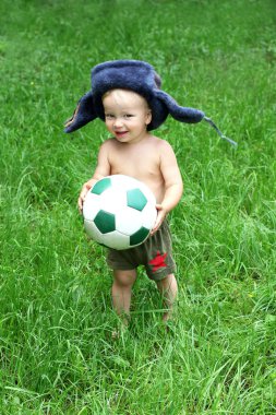 Ushanka şapkalı şirin Rus yürümeye başlayan çocuk futbol ya da futbol topu elinde tutuyor. Rusya futbol kavramı. Yeşil çim arka plan.