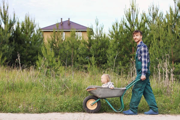 手押し車で家の近くの田舎道の息子を押す作業服の父 園芸の家族の概念 — ストック写真