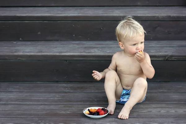 Cute toddler boy sitting on porch and eat fresh berries and fruits. Healthy lifestyle and happy childhood.