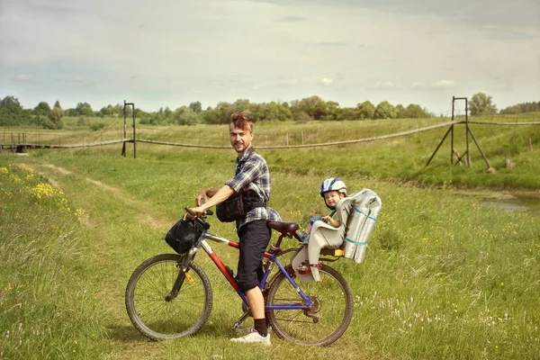 田舎で自転車旅行 自転車で父と赤ちゃん用の椅子に彼の幼児の息子 Suspenced 橋の近くの田舎道での走行します アクティブな家族概念 — ストック写真