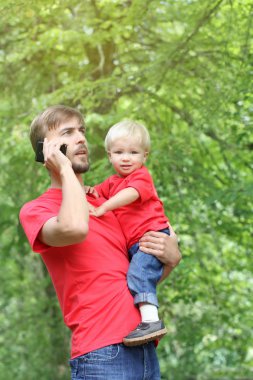 Babası yürümeye başlayan çocuk oğlu kollarını ve görüşmeler bir smartphone içinde tutar. Baba ve bebek açık. Aile giyim bak. Kopya spase. 