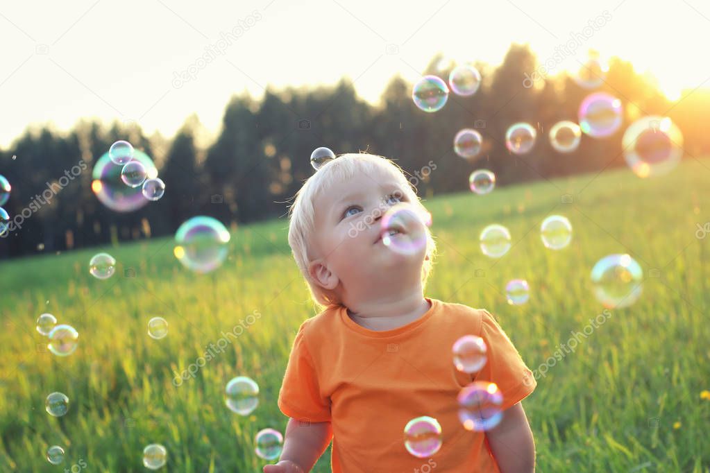 Cute toddler blond boy playing with soap bubbles on summer field. One bubble on his head. Happy childhood concept. Authentic lifestyle image.