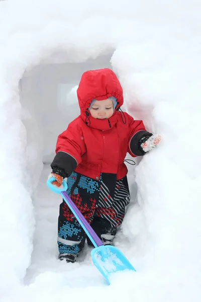 シャベルとかわいい男の子は 大きな雪の吹きだまりの雪の洞窟を掘っていきます 冬の楽しみの概念 — ストック写真