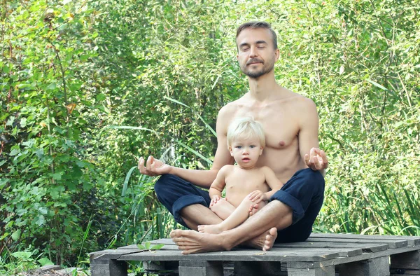 Father sitting in pose of lotus with their eyes closed and meditating, his little son is sitting on his lap. Family yoga concept.