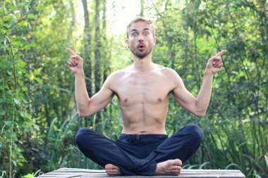 Genç adam oturur poz lotus açık ve noktası parmak sağ ve sol yönlere tarafından komik sakallı. Zemin üzerine yeşil orman.