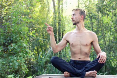 Genç Lotus açık poz oturan adam sakallı. Parmağını left.by için puan. Yeşil orman arka plan üzerinde. Metin için boş alan.