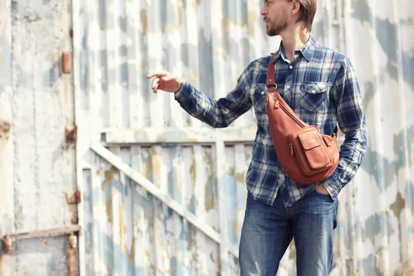 Şık deri sırt çantası ile rahat kıyafet genç hipster adam sağ elini tarafından işaret eder. Grunge duvar bir arka plan üzerinde. Modern kent yaşam tarzı. Çanta üzerinde odaklanmak.